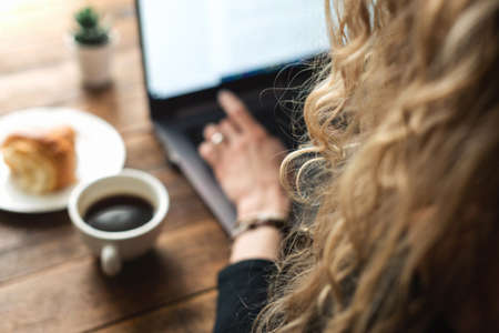 Unrecognizable blonde curly business woman working at laptop. Woman writing a book. Cup of coffee and croissant on sideの写真素材