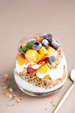 Greek yogurt, granola and fruits in jar. Healthy vegetarian breakfast parfait with peach, blueberries, oat honey granola and yogurt, beige backgroundの写真素材
