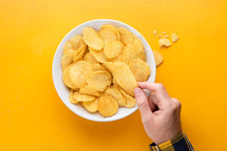 Spicy potato crisps in a bowl on yellow background. Male hand picking potato crisps. Junk food, unhealthy eating, party food conceptの写真素材