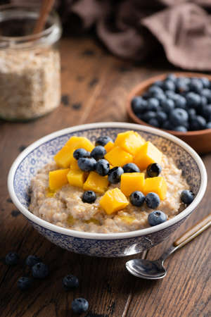 Oatmeal bowl with blueberries and mango on wooden table, healthy breakfast foodの写真素材