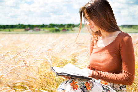 Fashion beautiful girl in the field with yellow spikelets on the sky background with bookの写真素材
