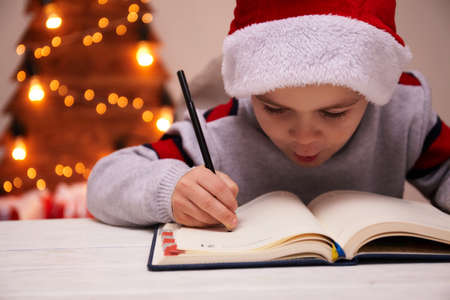 Christmas Child Write Letter to Santa Claus, Kid in Santa Hat Writing Wish List, unfocused lights backgroundの写真素材