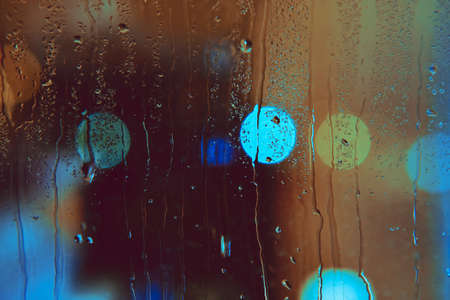 Rain drops run down the window with colorful lights of the night city. Abstract background of city life in night in rainy season.の写真素材