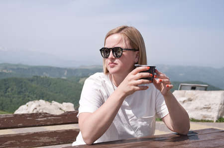 Woman in white t-shirt and black sunglasses drinking warm tea in the rustic wooden outdoor cafe on mountain area.の写真素材