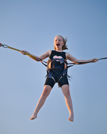 Girl child jumping on a trampoline with insurance elastic bands in an amusement park and carousels.
