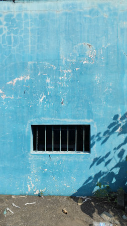 A weathered bright blue wall with barred windows and peeling paint details