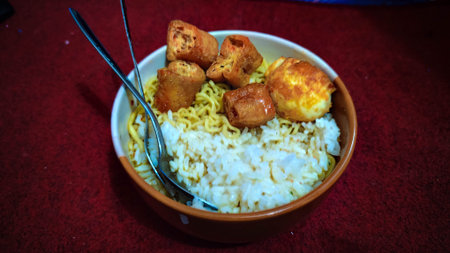 An overhead view of an Indonesian food bowl containing cooked rice, instant noodles, fried tofu/tempeh pieces, and a fried egg, with metal utensils resting insideの写真素材