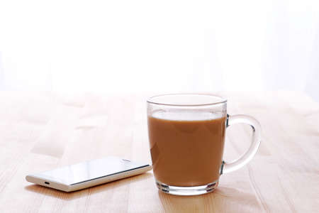 glass coffee with milk mug and smartphone lit by morning sunlight on a wooden table, copy spaceの写真素材