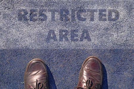 man feet in brown leather shoes standing before shabby sign restricted area written on asphalt, top viewの写真素材