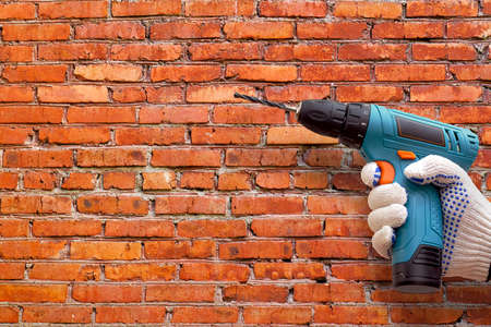 hand in dotted work glove holding cordless drill machine on brick wall background, copy spaceの写真素材
