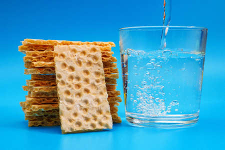 Pile of rye crispbread and water pouring into glass on blue background