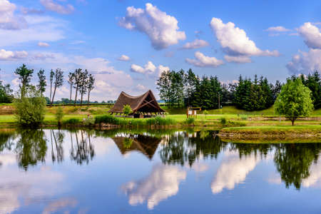 A small traditional cottage covered with straw at a lake, beautiful reflection of clouds in the water, at the far shore people are fishing (face not recognizable)の写真素材