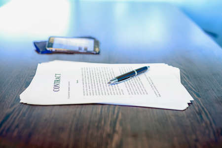 Business contract / agreement and fountain pen on dark wooden desk, with mobile phone in background, shallow depth of field, low angleの写真素材