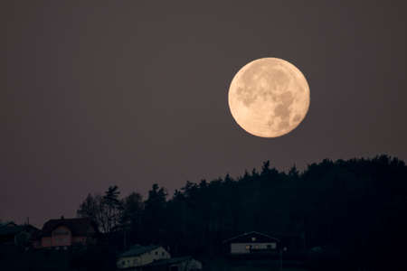 Setting full moon behind hill, forest and houses seen on hillの写真素材