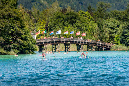 Portschach, Austria - August 14 2016: International flags flying over the reiling of the wooden bridge leading from Portschach to the Blumeninsel (flower island) while swimmers enjoy the water on a hot summer day.の写真素材