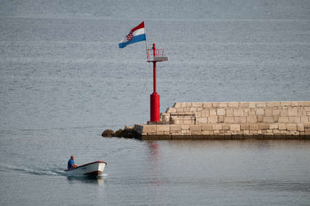 Vinjerac, Croatia - July 28 2018: Local fisherman in small boat entering harbor coming into town for basic supplies needed for living in remote locations. Croatian flag flying high from lighthouseのeditorial素材