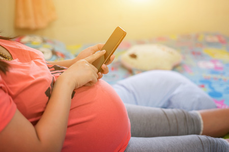 pregnancy, motherhood, technology, people and expectation concept - close up of pregnant woman with smartphone in bed at home, using mobile phone in her bedroomの写真素材