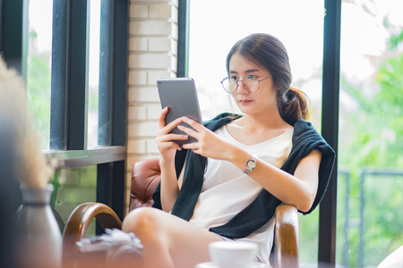 Young asian female entrepreneur reading electronic book on digital tablet during in modern restaurant interior, elegant businesswoman chatting on touch pad while sitting in comfortable coffee shopの写真素材
