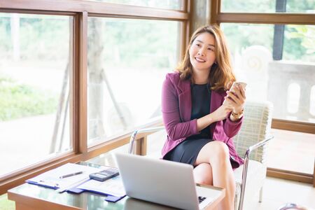 Smiling Asian woman sitting and using a smart phone in the cafe, Beautiful young Asian girl working at a coffee shop with a laptop.female freelancer connecting to internet, business conceptの写真素材