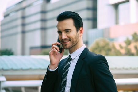 Handsome young businessman standing outdoors and talking with partnership by smartphone,using mobile phone control the business in daily life,internet of things IOT ,Business concept. の写真素材