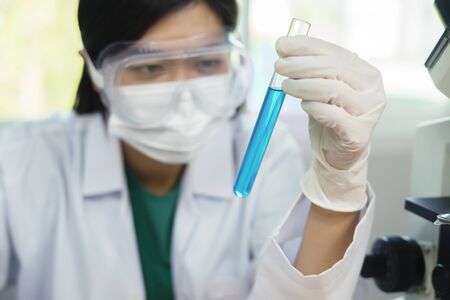 Asian Female medical Doctor or research scientist looking at a test tube of clear solution in a lab or laboratory with microscope and micro cell slide,chemistry, technology, biology people conceptの写真素材