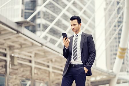 Handsome young businessman standing outdoors and looking at smartphone,using mobile phone control the business in daily life,internet of things IOT ,Business concept.の写真素材