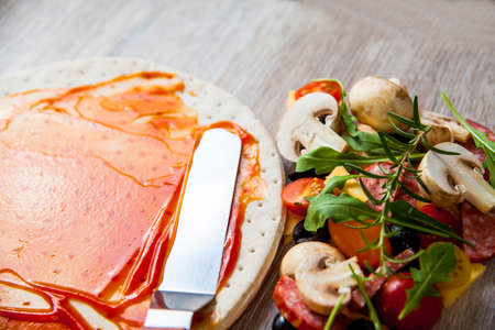 Dough basis covered with ketchup and ingredients for pizza. On a wooden table.の写真素材