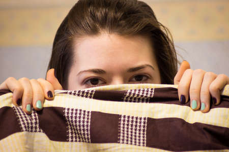 Shy cute lovely young woman with lying in bed and hiding her faceの写真素材