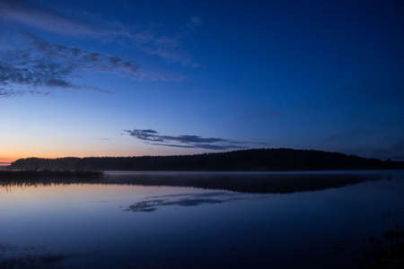 beginning sunrise on a big lake with forests and grass with beautiful mist and cloudsの写真素材
