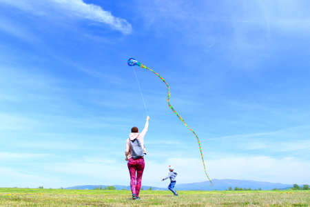 Happy family, mom and son, playing with flying kite on meadow in beautiful spring sunny day, blue sky in backgroundの写真素材