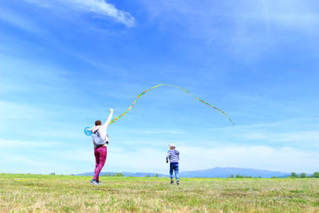 Happy family, mom and son, playing with flying kite on meadow in beautiful spring sunny day, blue sky in backgroundの写真素材