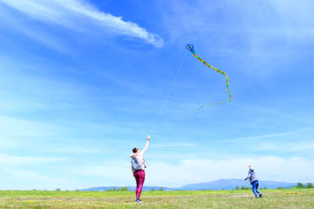 Happy family, mom and son, playing with flying kite on meadow in beautiful spring sunny day, blue sky in backgroundの写真素材