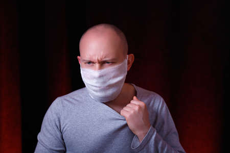 A frowned man with emotion of pain in medical gauze mask standing on dark red background. Protection of upper and lower respiratory tract from coronavirus. Virus preventive action concept.の写真素材