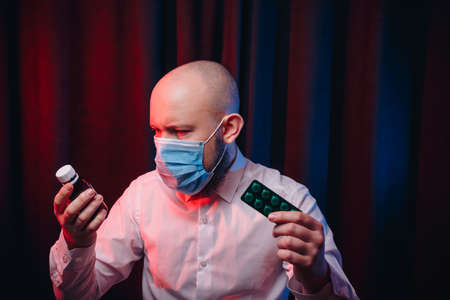 Frowned man in gauze mask and white shirt holding medicines in hands on dark background. Healthcare, respiratory illness prevention, prophylaxis of virus infections, COVID-19 concept.の写真素材