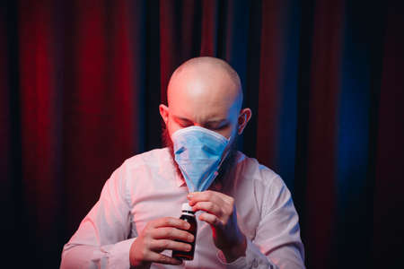 Frowned man in gauze mask and white shirt holding medicines in hands and trying to drink mixture on dark background. Healthcare, respiratory illness prevention, prophylaxis of virus infections, COVID-19 concept.の写真素材