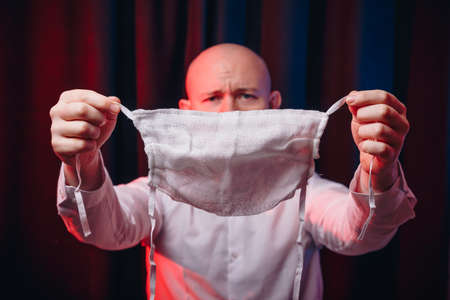 Frowned man in white shirt holding gauze mask in hands on dark background. Emotion of pain, fear and...