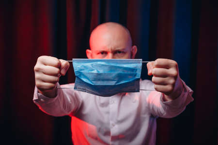 Frowned man in white shirt holding gauze mask in hands on dark background. Emotion of anger and determination. Healthcare, respiratory illness prevention, prophylaxis of virus infections, COVID-19 concept.の写真素材