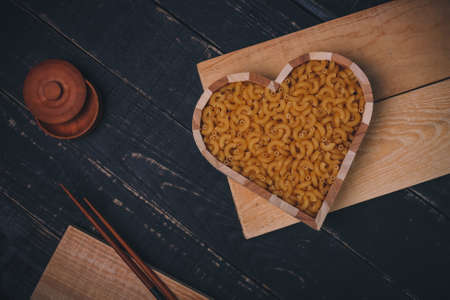 Wooden heart shaped box with pasta or raw macaroni, salt pot and chopsticks on black wooden background, copy space. Carbohydrates diet, healthy eating habits and vegetarian food concept.の写真素材