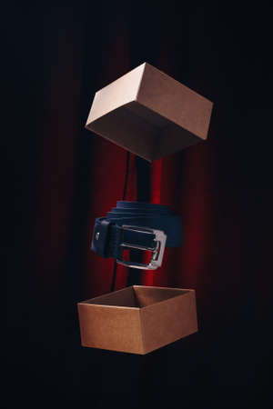 Levitating leather belt or waistband and carton box with cover on dark background with red light. Male accessories concept, vertical shot.の写真素材