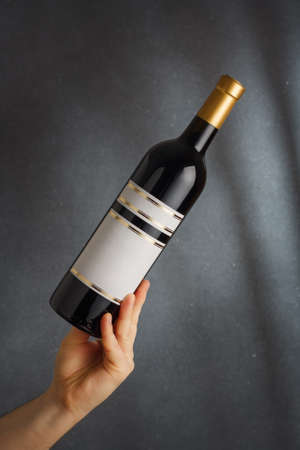 Male hand holding bottle of red wine, natural fruit alcohol liquor with empty label on gray background. Mock up, template for brand, vertical shotの写真素材