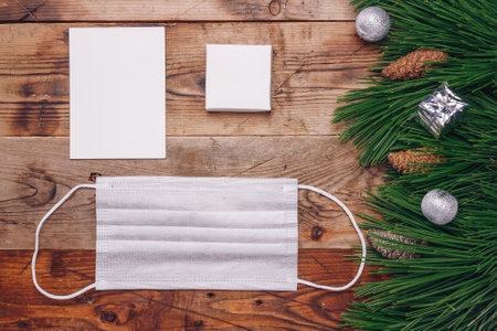 Safe Christmas . Pandemic New Year 2021 concept. Pine branches, fir cones, paper template mock up and protective gauze mask on wooden brown background.の写真素材