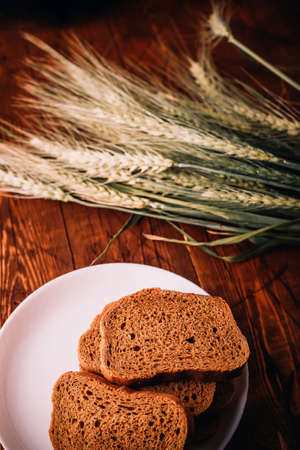 Slices of rye wholegrain bread on white plate, wheat ears on wooden boards background, rustic kitchen table. Homemade bread, bakingの写真素材