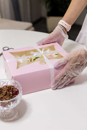 confectioner holding pink box of cupcakes, hands in gloves close. Vase of dried rose petals on table. Mother's day, Valentine's day, birthdayの写真素材