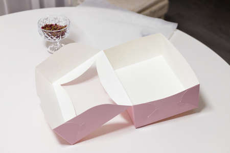 Pink empty paper box for sweet cakes, cupcakes or muffins on white kitchen table. Vase of dried rose...