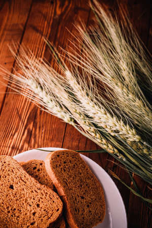Slices of rye wholegrain bread on white plate, wheat ears on wooden boards background, rustic...