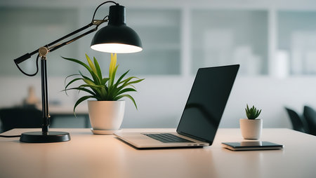 Laptop on a desk in a bright office with a lamp and a plantの素材