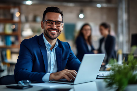 Portrait of smiling businessman working on laptop in office. Business people in the background.の素材