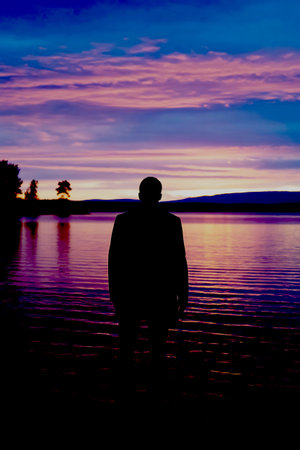 A silhouette of a man standing on the shore of a lake at sunsetの写真素材