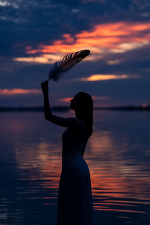 Silhouette of a beautiful woman in a white dress with a feather in her hand against the background of a beautiful sunsetの写真素材