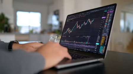 A person sits at a desk, intently studying financial data on a laptop screen displaying various graphs and charts, with a blurred background of a modern room.の写真素材